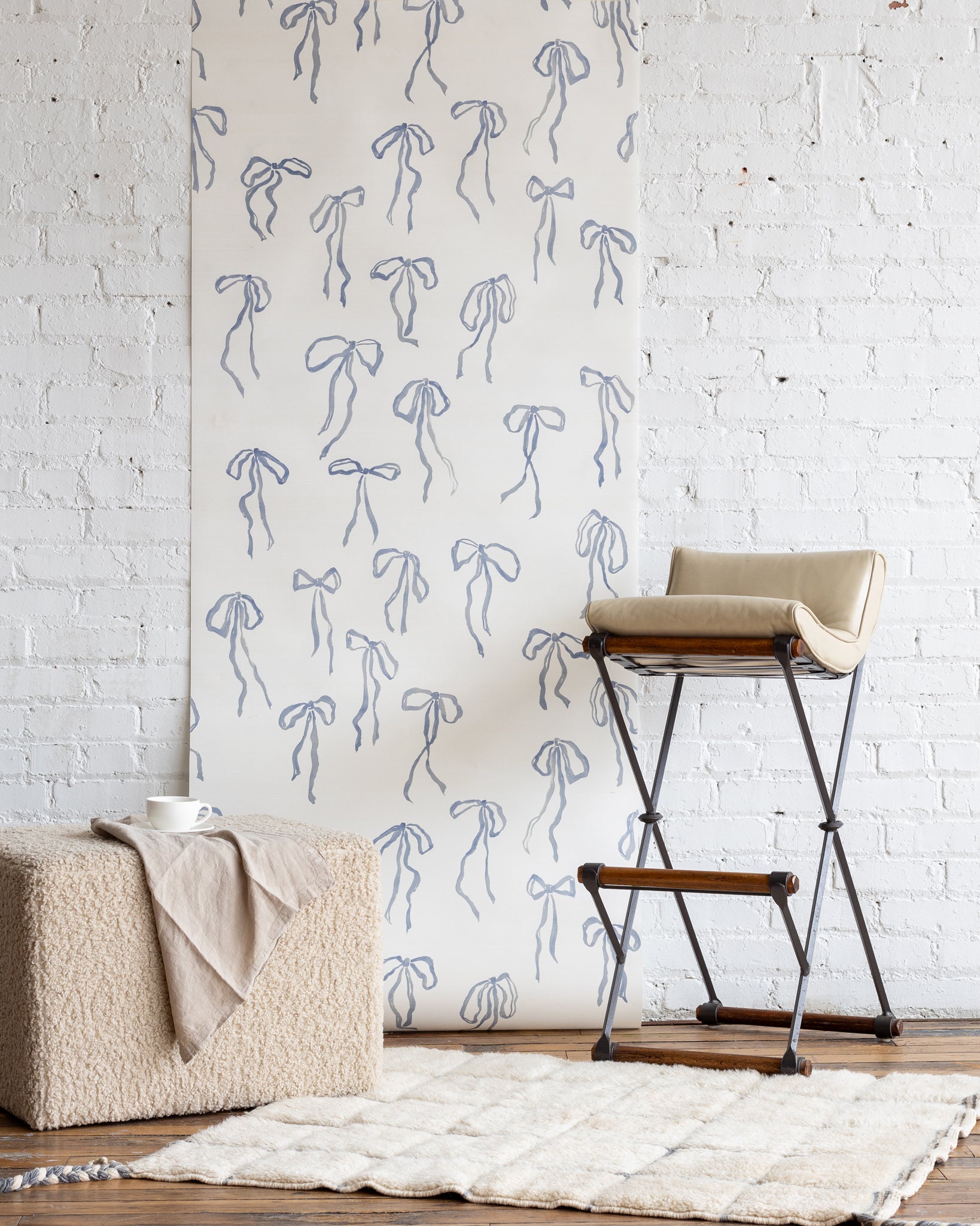 Elegant silk wallpaper featuring hand-drawn blue bows on an off-white background, complemented by a boucle ottoman, a beige barstool, and a plush white rug.