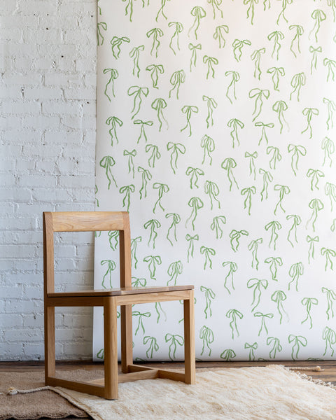 Playful wallpaper with hand-drawn green bows on a white background, styled with a natural wood chair and a textured cream rug.