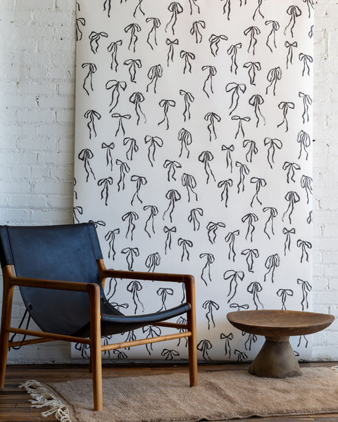 Minimalist wallpaper featuring black hand-drawn bows on a white background, styled with a black leather and wood lounge chair in a rustic interior.