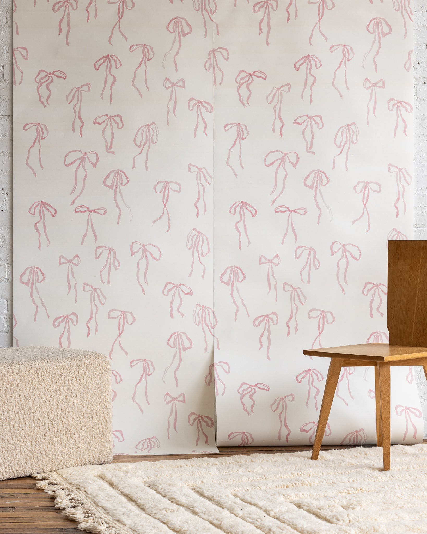 Feminine wallpaper with delicate pink hand-drawn bows on a white background, styled with a boucle ottoman, wooden chair, and a plush cream rug.