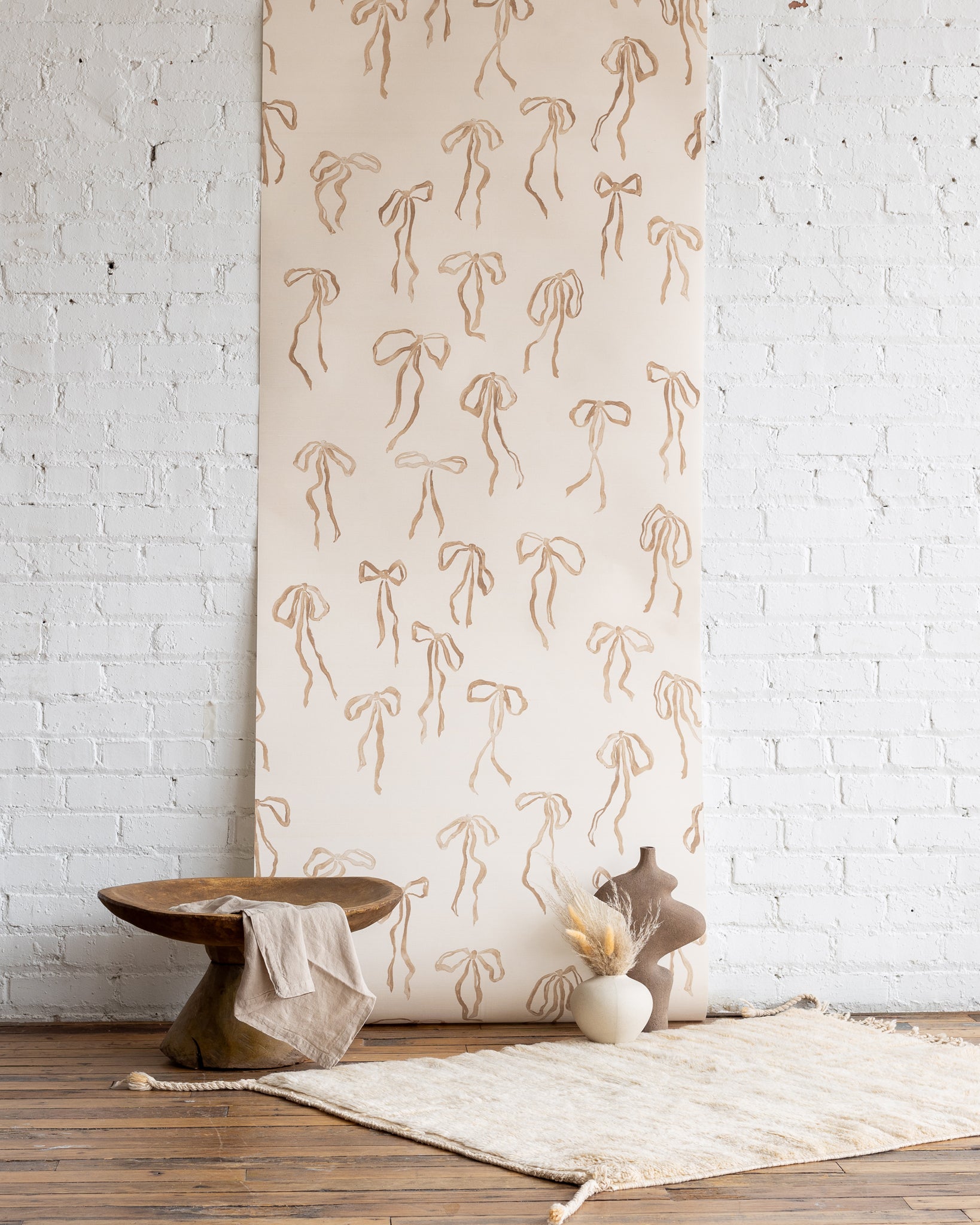 Neutral-toned wallpaper with hand-drawn brown bows on a warm beige background, styled with a rustic wooden table, sculptural vase, and soft natural decor.