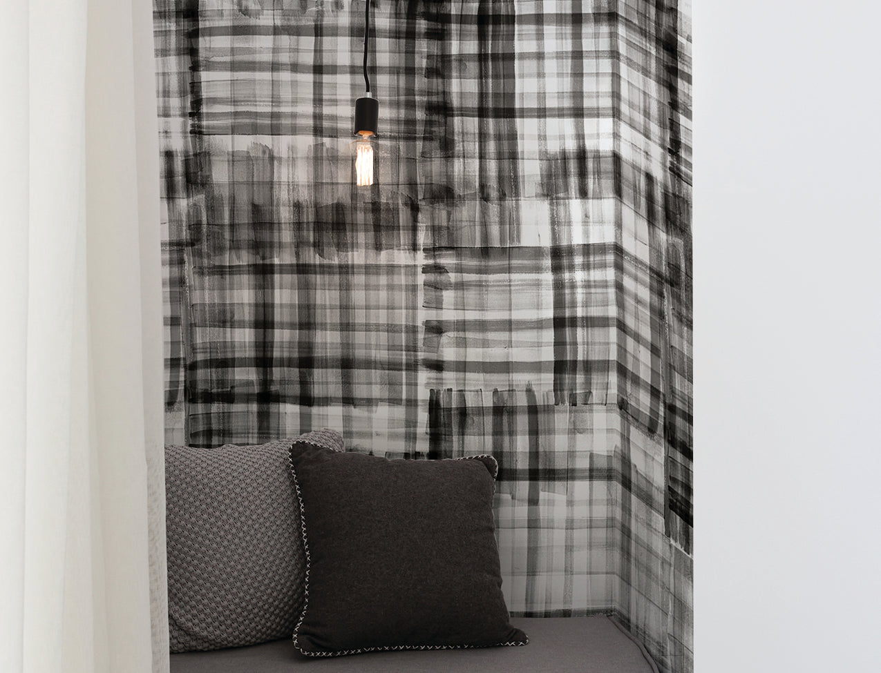 Cozy nook with black and white hand-painted plaid wallpaper, complemented by a minimalist pendant light, dark gray cushions, and soft drapery.