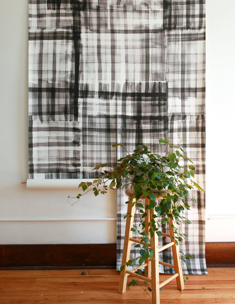 Modern interior with a roll of black and white watercolor plaid wallpaper partially unrolled against a white wall, styled with a wooden stool and cascading green plant.