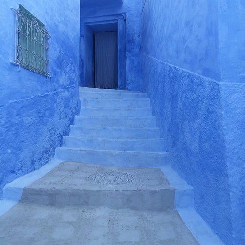 Narrow stairway leading to a doorway, surrounded by vibrant blue-washed walls in Chefchaouen, Morocco, with a decorative metal window grille on the left.