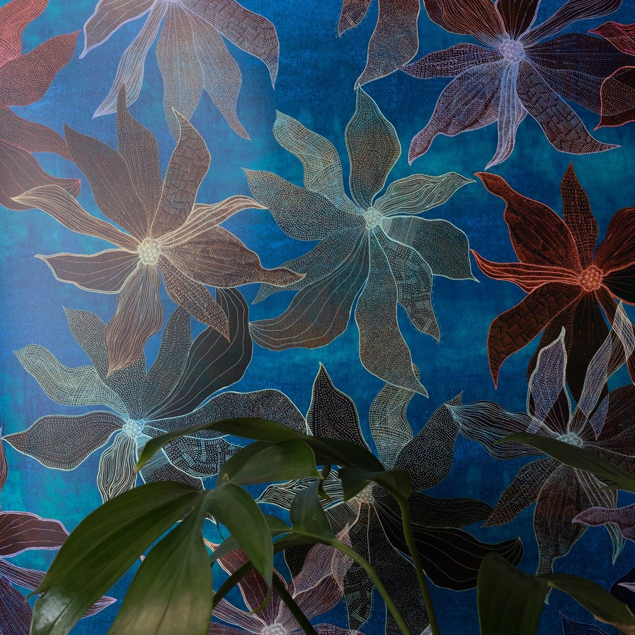 Blue floral wallpaper pattern featuring intricate line details; green houseplant leaves in the foreground adding a natural element.
