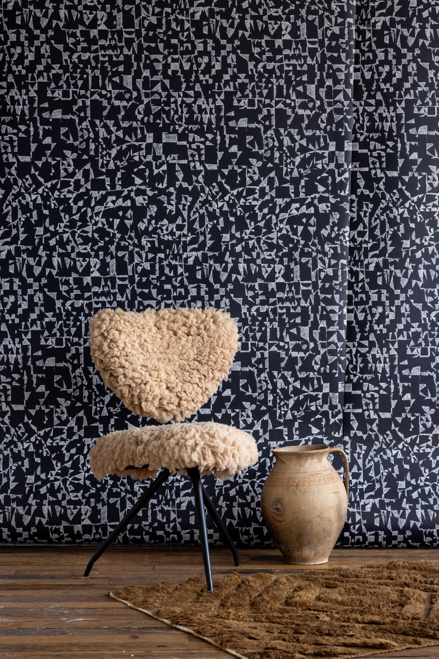Black wallpaper with intricate white geometric shapes, styled with a beige shearling chair, rustic clay jug, and textured brown rug on wooden floor.