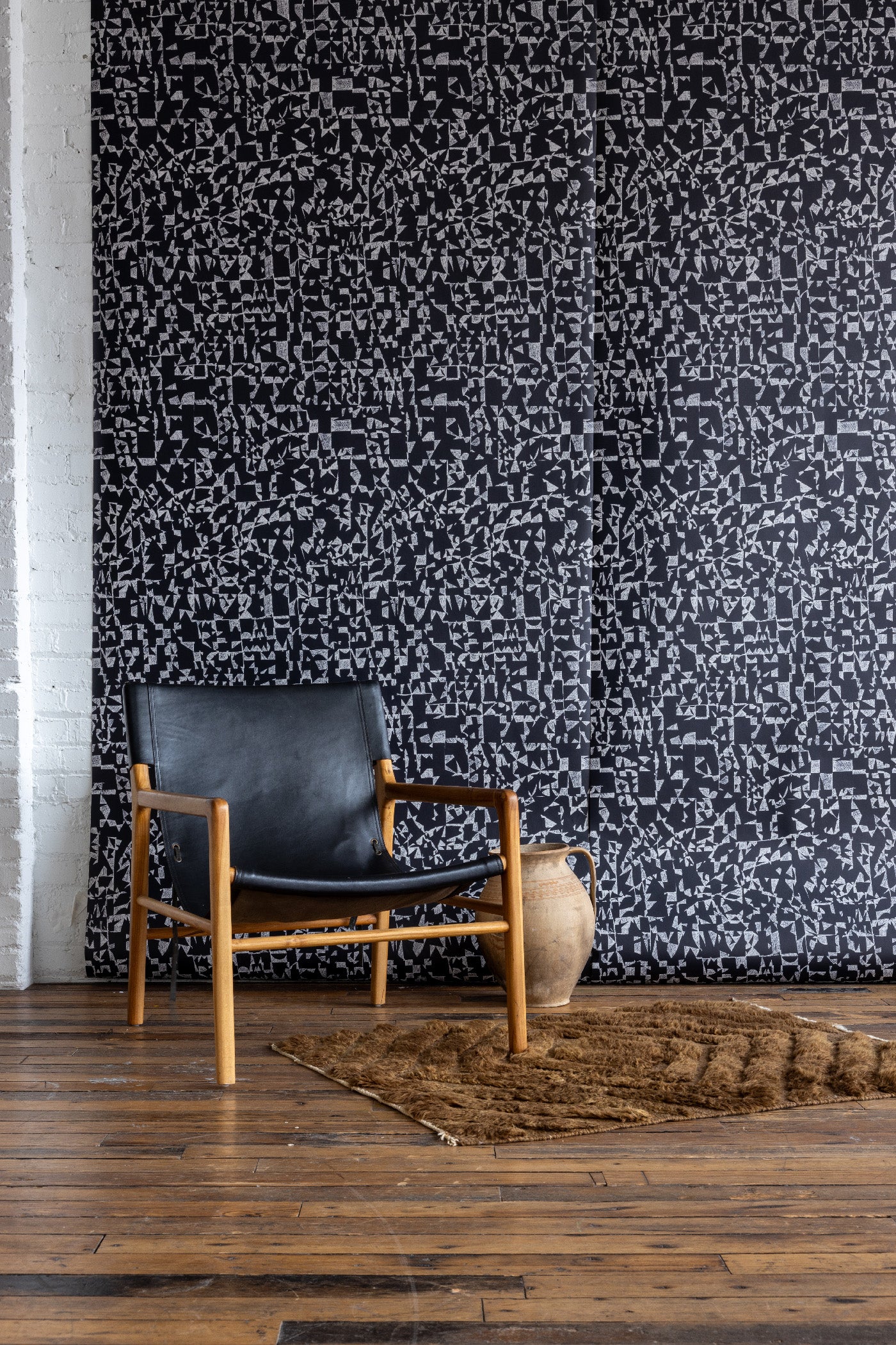 Black wallpaper featuring white abstract geometric patterns, paired with a black leather and wood chair, clay jug, and brown textured rug on wooden floor.
