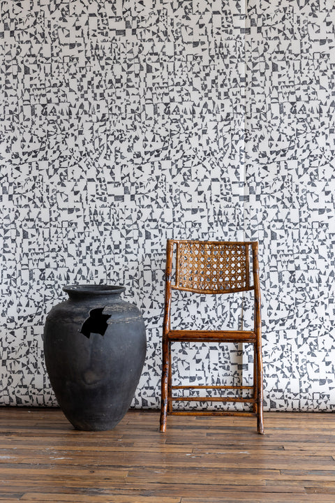 White wallpaper with black geometric pattern, styled with a black clay pot and a woven cane chair on wooden floor.
