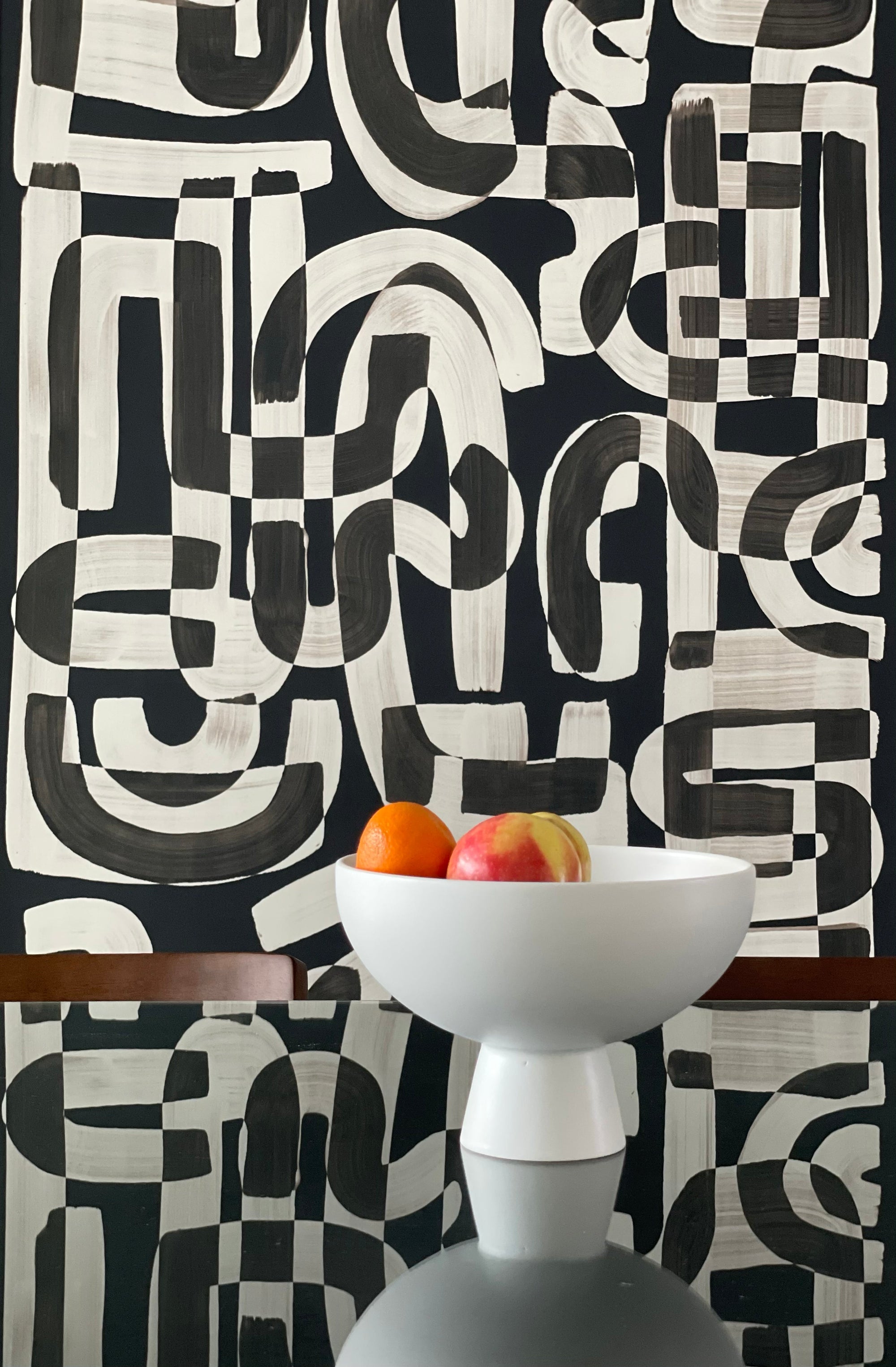 Black and white abstract wallpaper called Down the Rabbit Hole in a dining room setting, with a modern white bowl filled with oranges and an apple placed on a reflective glass table.
