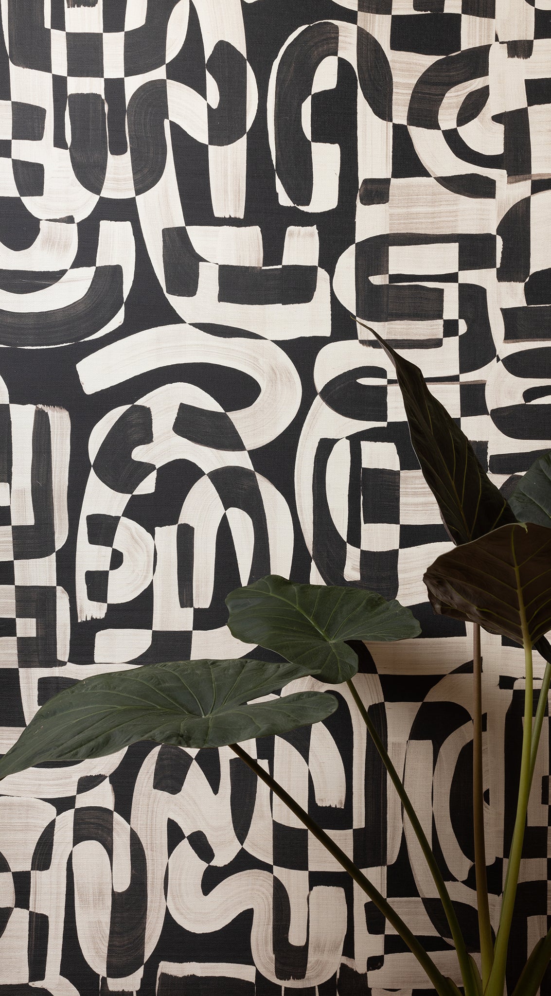 Close-up of black and white grasscloth wallpaper with bold, abstract brushstroke patterns, partially obscured by large green tropical leaves.