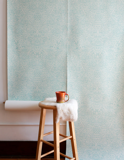 Light blue wallpaper with an intricate feather-like pattern displayed against a white wall, with a wooden stool draped in faux fur and a copper mug on top.