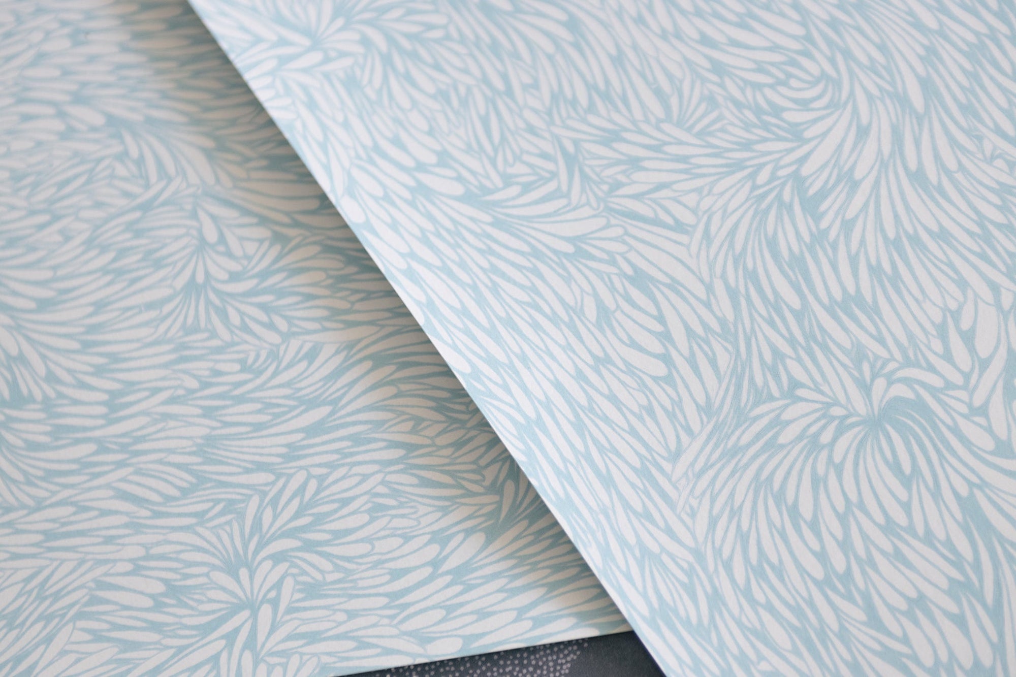 Overlapping wallpaper samples with a soft blue and white intricate feather-like design, showcasing the texture and pattern.