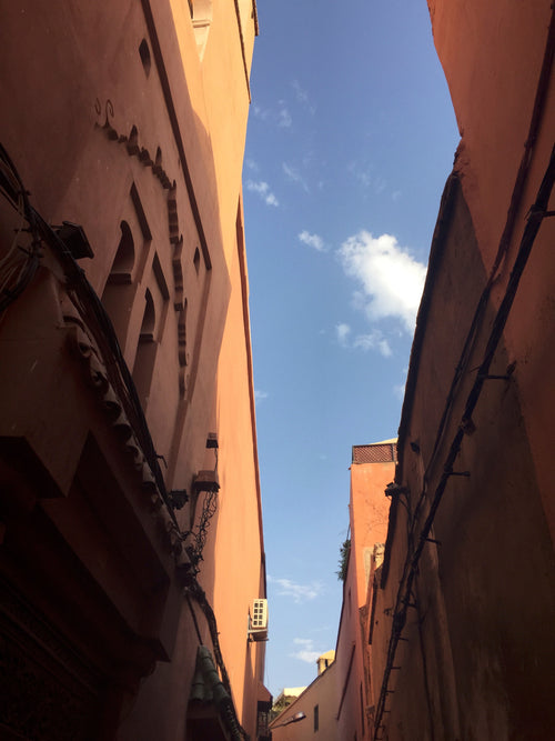 Narrow alleyway between traditional Moroccan buildings with warm, earthy-toned walls, intricate architectural details, and visible electrical wiring, leading up to a bright blue sky with scattered clouds.