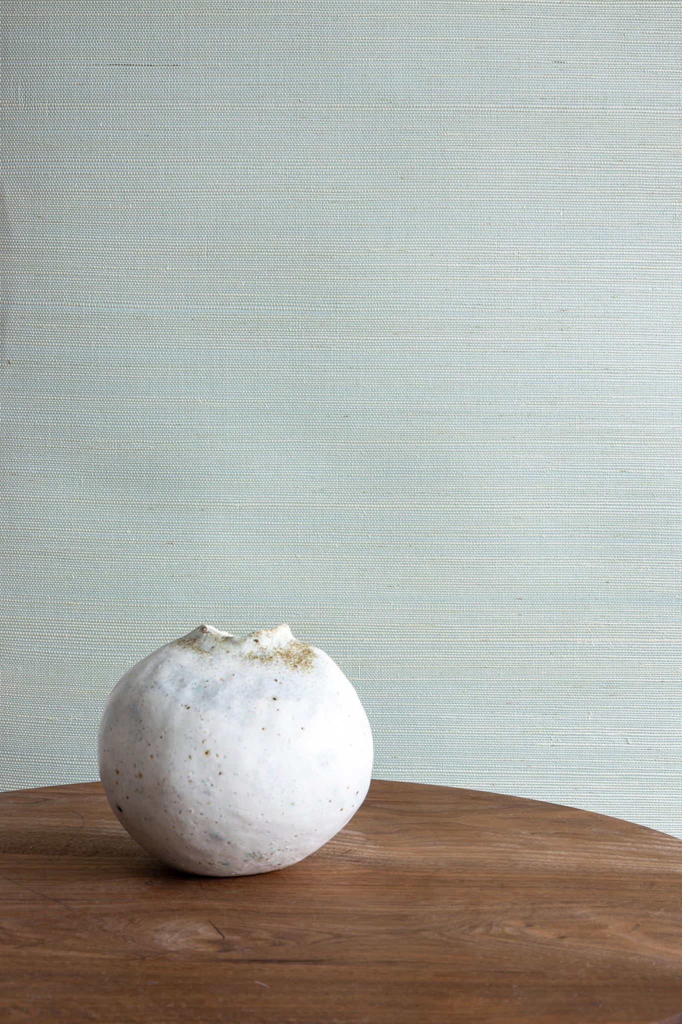 Soft light blue textured wallpaper with a woven finish, styled with a wooden side table and a single white ceramic vase.