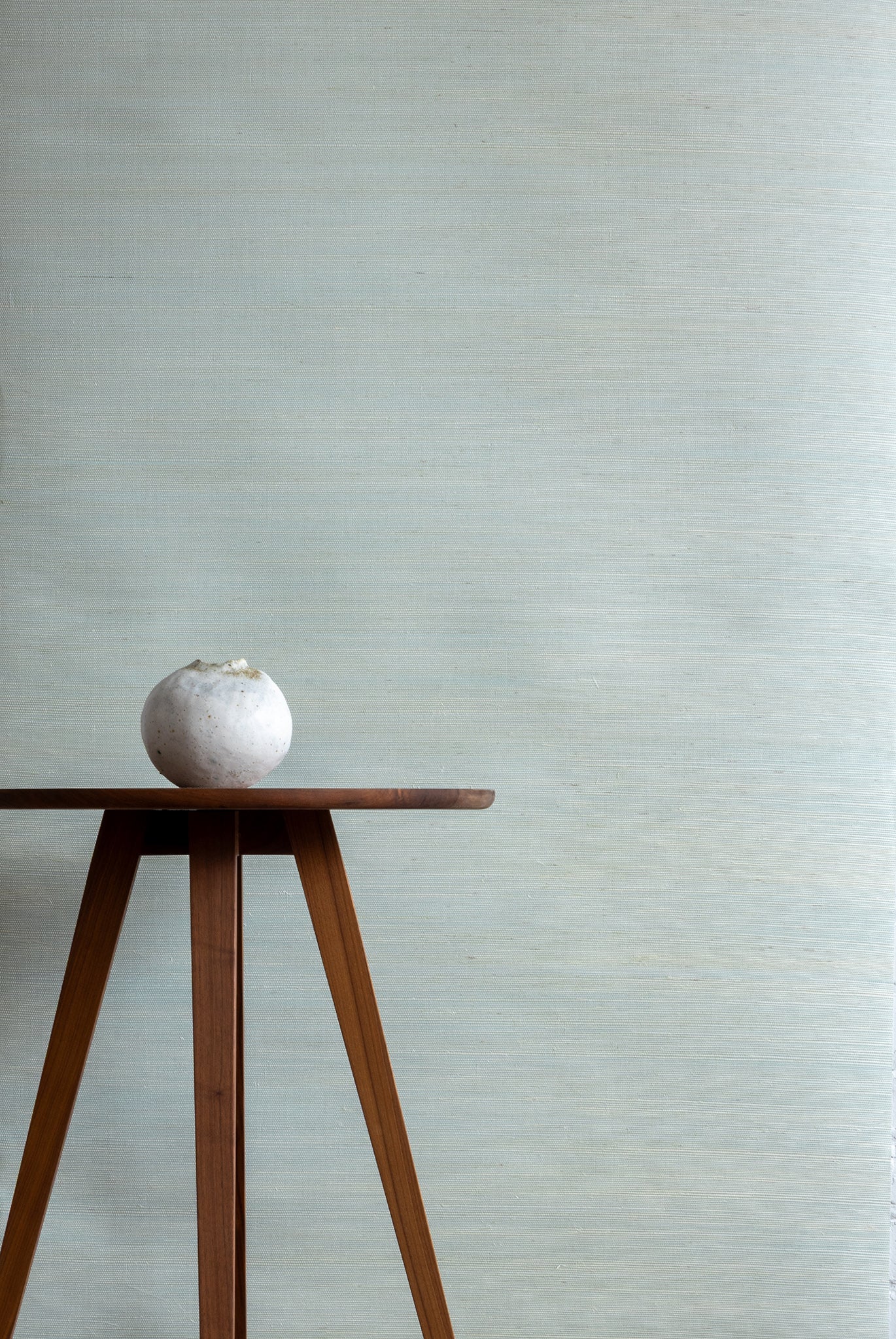 Soft pale blue textured grasscloth wallpaper with a subtle woven finish, styled with a wooden tripod side table and a small white ceramic vase.