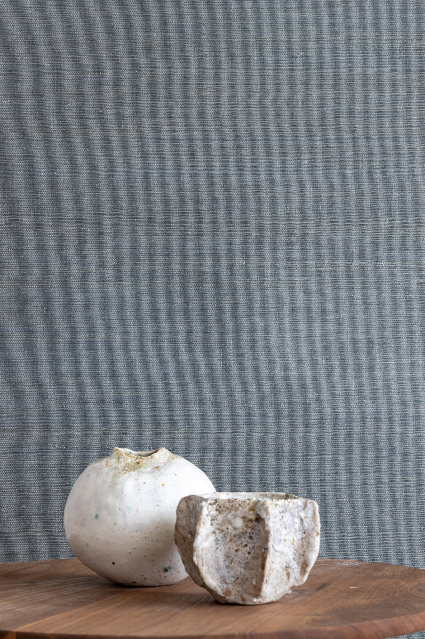 Blue textured grasscloth wallpaper with a subtle woven finish, styled with two white ceramic vases on a wooden surface.