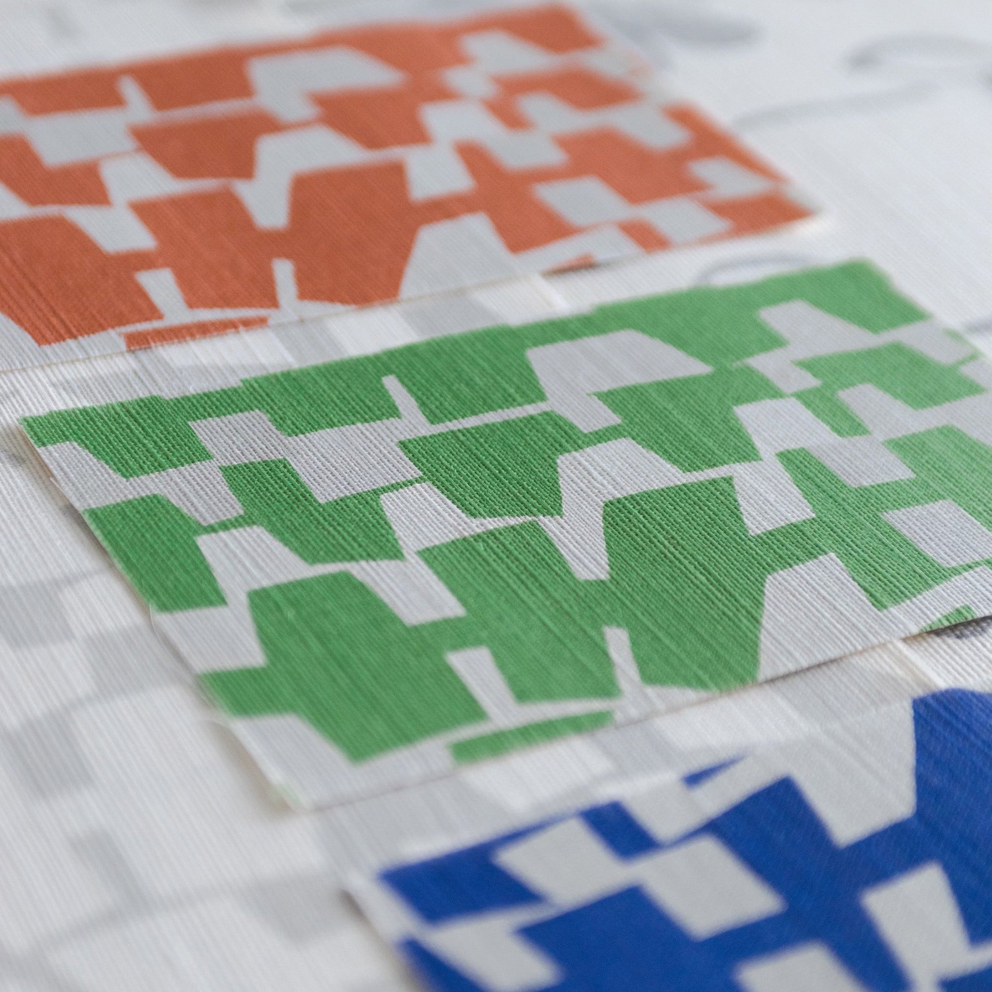 A grasscloth wallpaper sample sheet displaying three color variations of a geometric design—orange, green, and blue—on an off-white background.