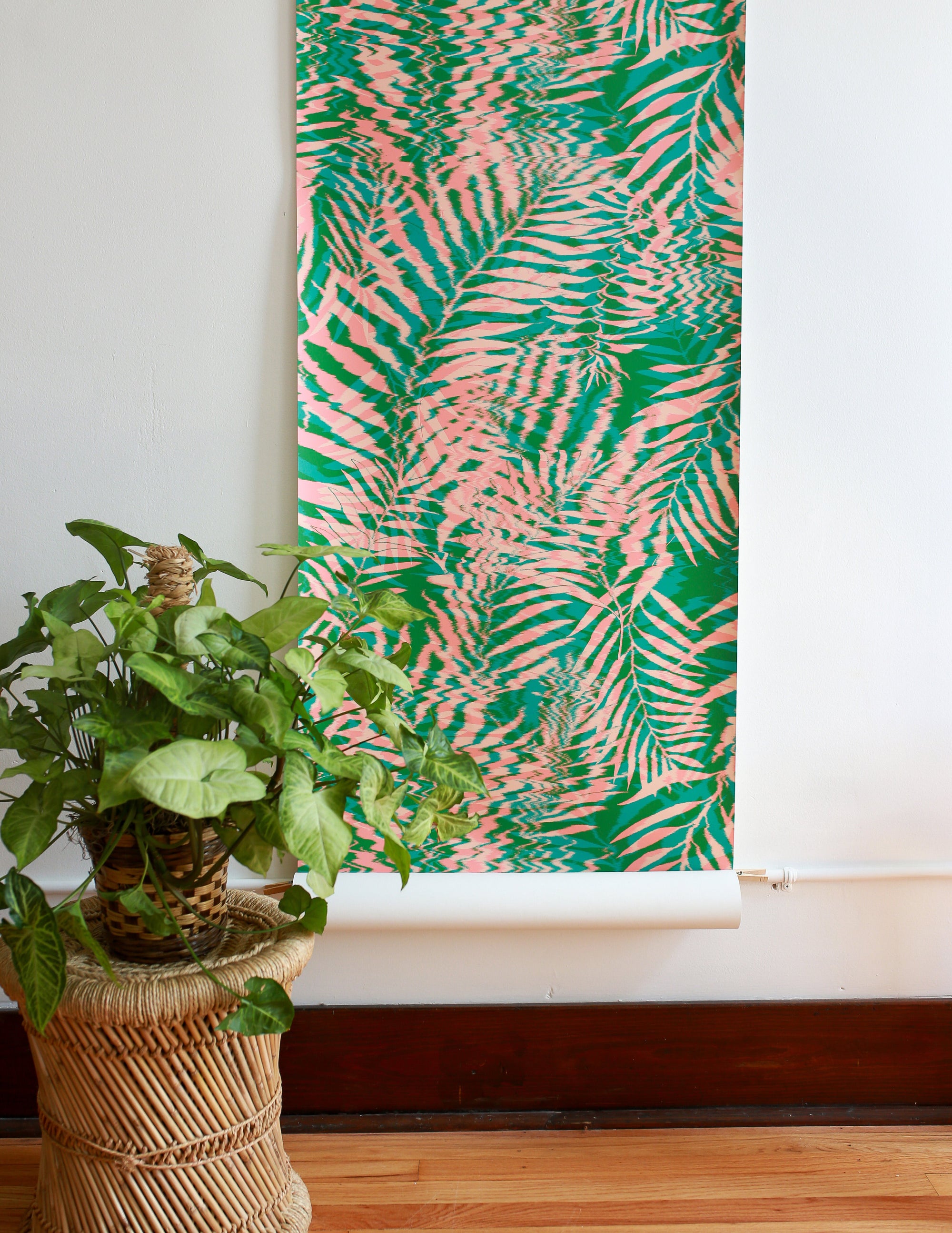 Tropical leaf wallpaper in vivid green and soft coral pink with a ripple effect, partially unrolled against a neutral wall next to a woven plant stand.