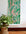 Tropical leaf wallpaper in vivid green and soft coral pink with a ripple effect, partially unrolled against a neutral wall next to a woven plant stand.