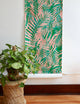 Tropical leaf wallpaper in vivid green and soft coral pink with a ripple effect, partially unrolled against a neutral wall next to a woven plant stand.