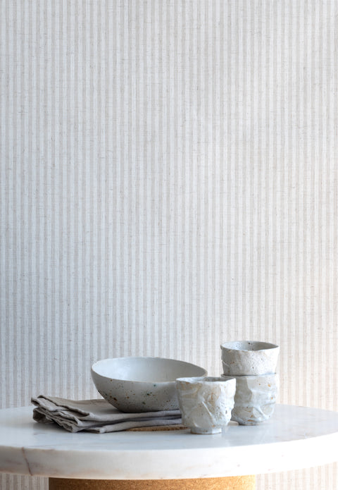 Interior setting featuring beige and ivory striped linen wallpaper behind a marble tabletop with ceramic dishware and folded linens.