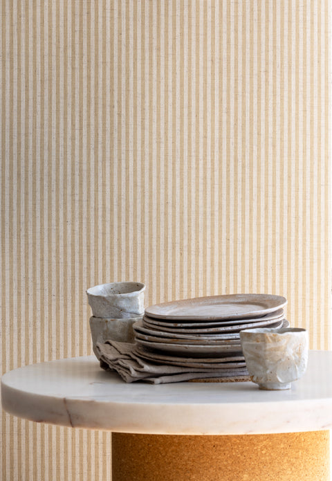 Interior setting with tan and off-white striped linen wallpaper as a backdrop, paired with stacked ceramic dishes and a marble tabletop.