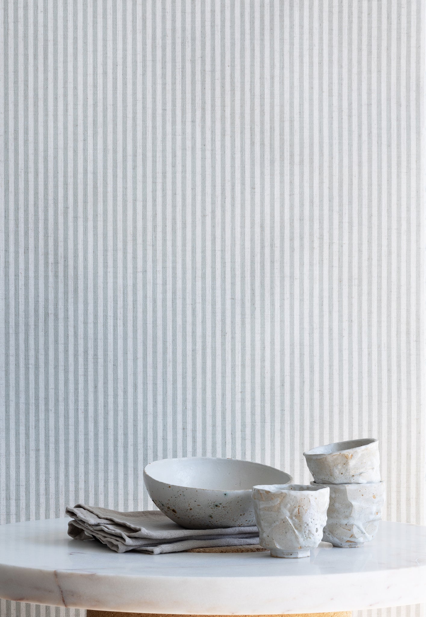 Styled interior scene with sage green and white striped linen wallpaper, paired with a marble tabletop, ceramic bowls, and neutral linens.