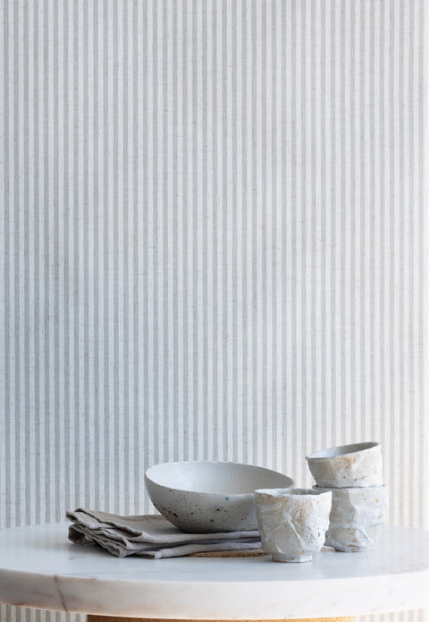 Styled interior scene with sage green and white striped linen wallpaper, paired with a marble tabletop, ceramic bowls, and neutral linens.