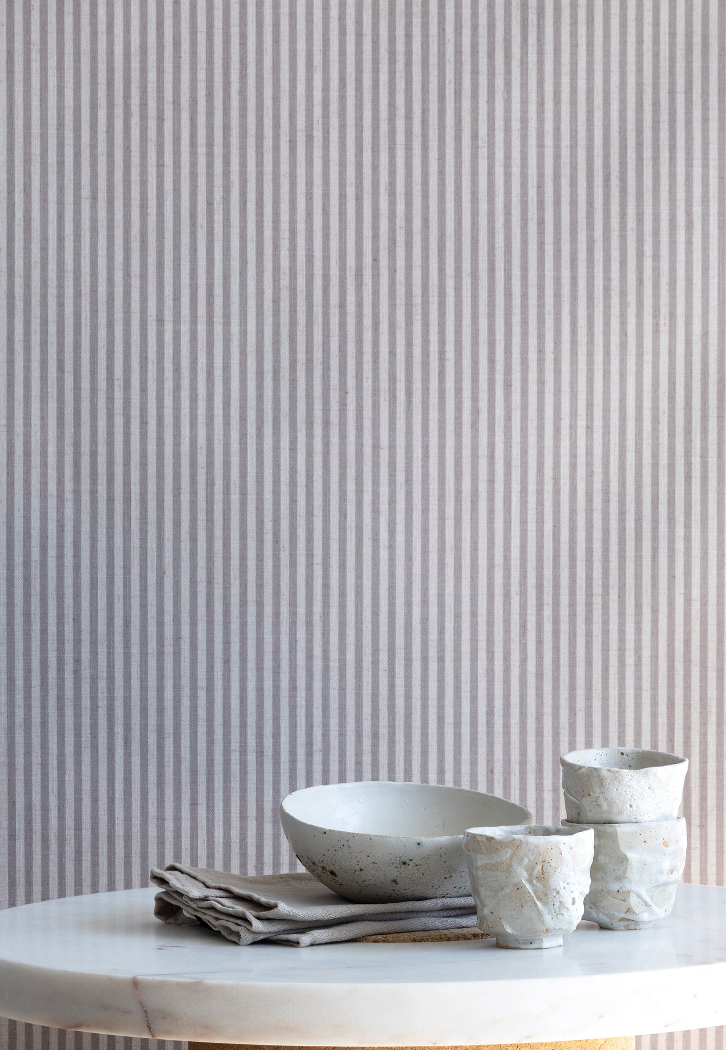 Interior scene with charcoal-striped linen wallpaper behind a marble table styled with handmade ceramic cups and a neutral linen napkin.
