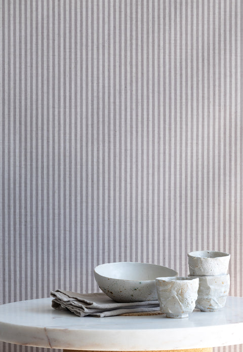 Interior scene with charcoal-striped linen wallpaper behind a marble table styled with handmade ceramic cups and a neutral linen napkin.