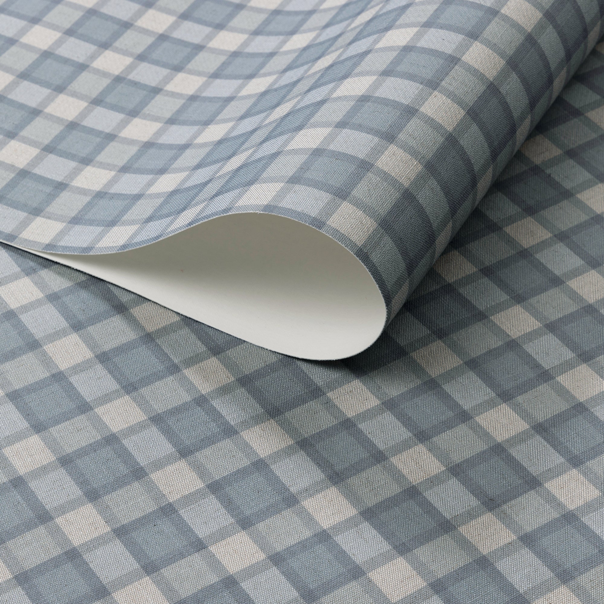 Close-up of vintage plaid check wallpaper in muted denim blue and cream tones with a soft linen texture, shown with a curled edge to display paper thickness.