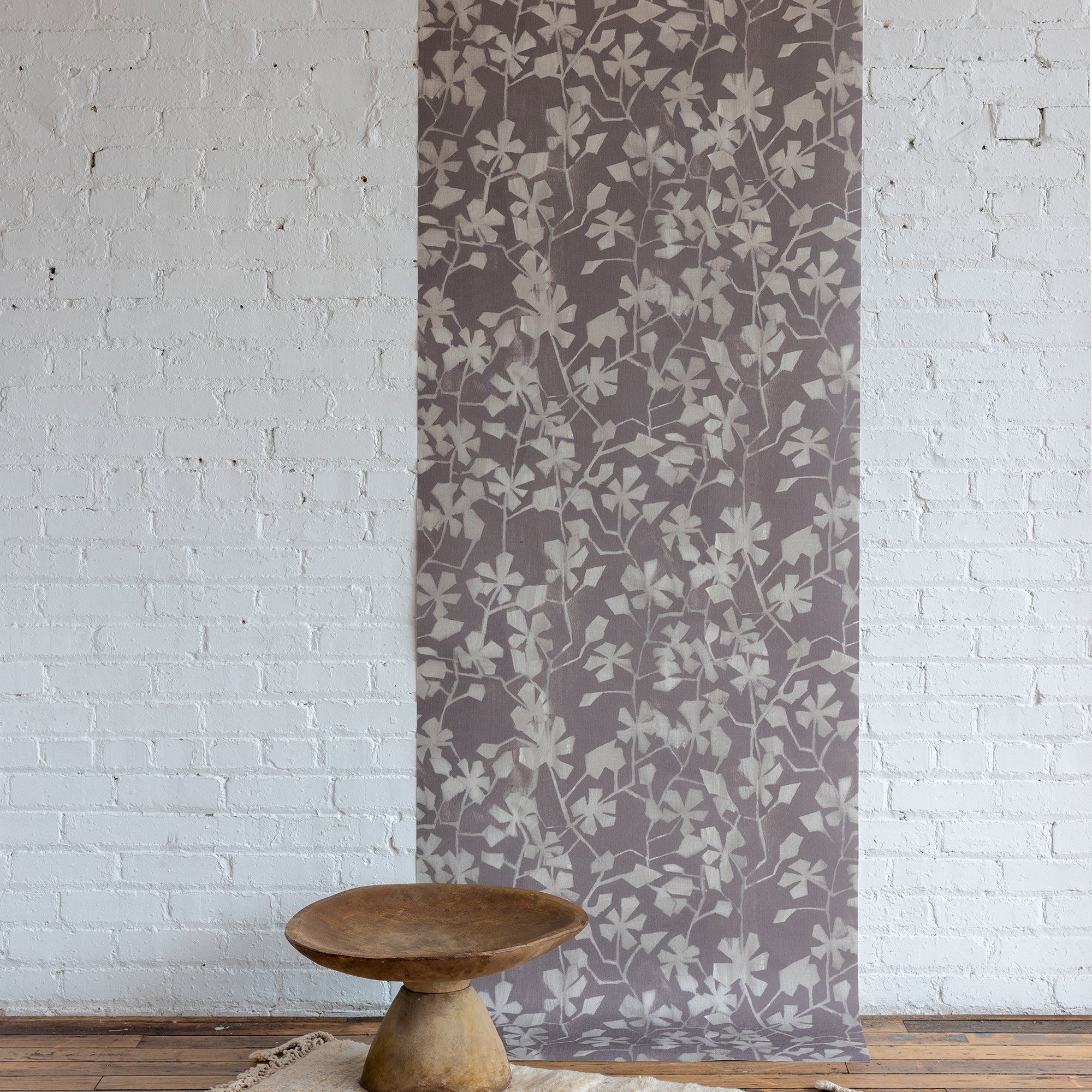 Soft botanical wallpaper featuring a muted mauve background with a subtle ivory leafy vine pattern, displayed against a white brick wall with a carved wooden pedestal bowl