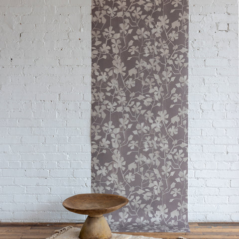 Soft botanical wallpaper featuring a muted mauve background with a subtle ivory leafy vine pattern, displayed against a white brick wall with a carved wooden pedestal bowl