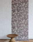 Soft botanical wallpaper featuring a muted mauve background with a subtle ivory leafy vine pattern, displayed against a white brick wall with a carved wooden pedestal bowl