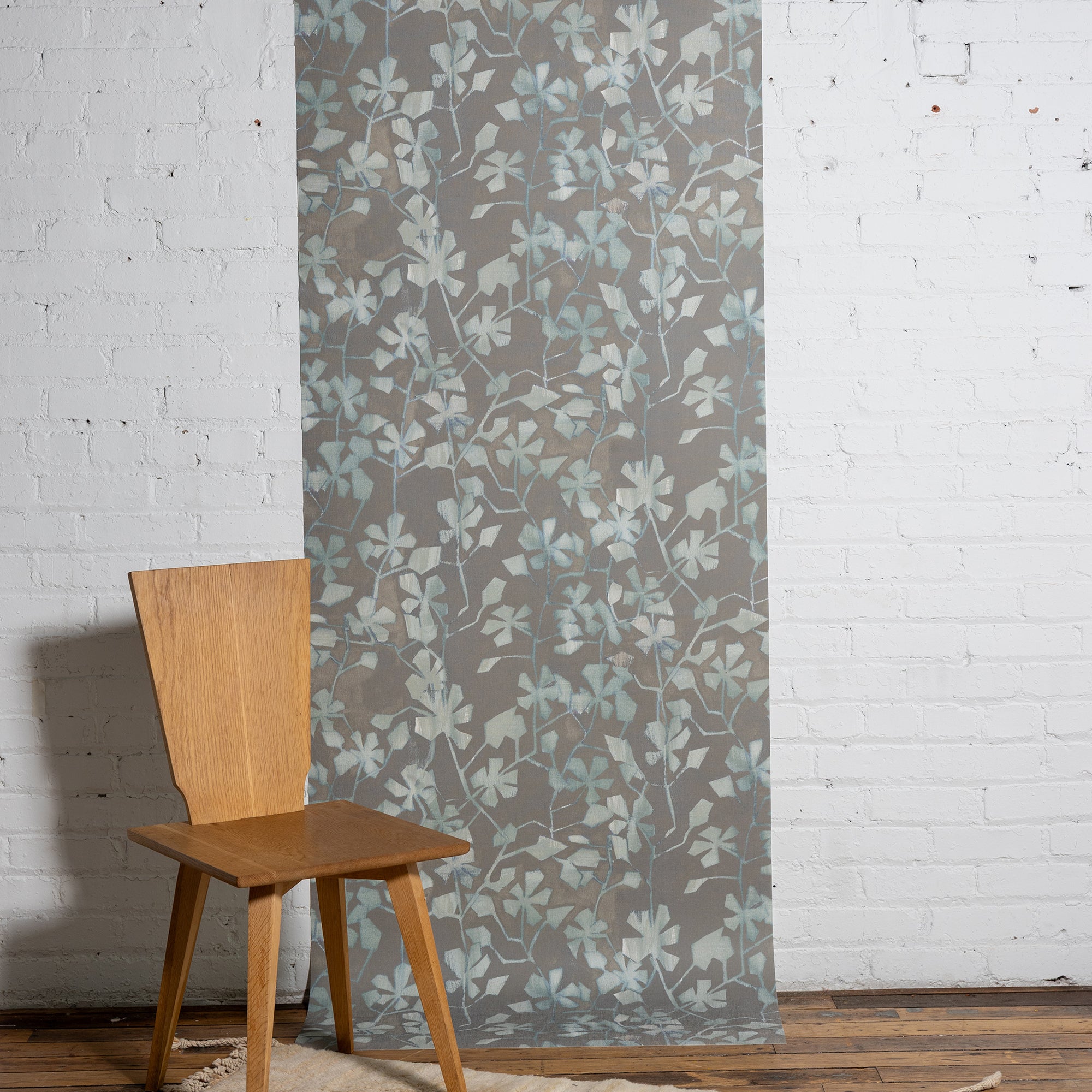 Botanical wallpaper with a taupe-gray background and light blue and ivory leafy vine pattern, styled against a white brick wall with a minimalist wooden chair.