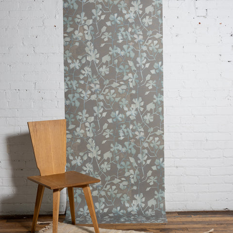 Botanical wallpaper with a taupe-gray background and light blue and ivory leafy vine pattern, styled against a white brick wall with a minimalist wooden chair.