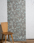 Botanical wallpaper with a taupe-gray background and light blue and ivory leafy vine pattern, styled against a white brick wall with a minimalist wooden chair.