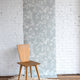 Light botanical wallpaper with a soft blue-gray background and white leafy vine pattern, shown against a white brick wall with a natural wood chair on a neutral woven rug.