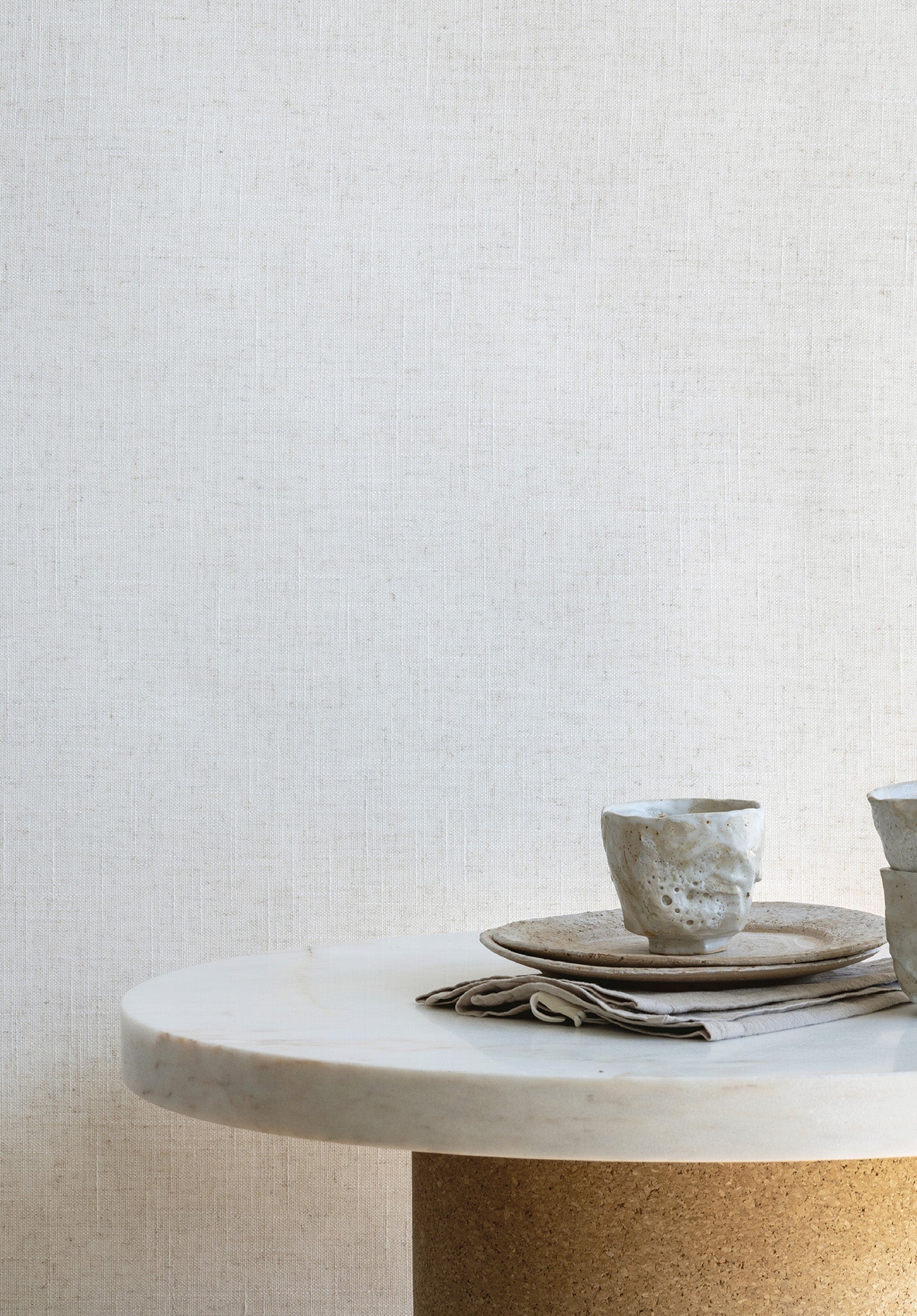 Neutral-toned linen wallpaper with a subtle woven linen texture, styled with ceramic tableware on a marble and cork pedestal table.