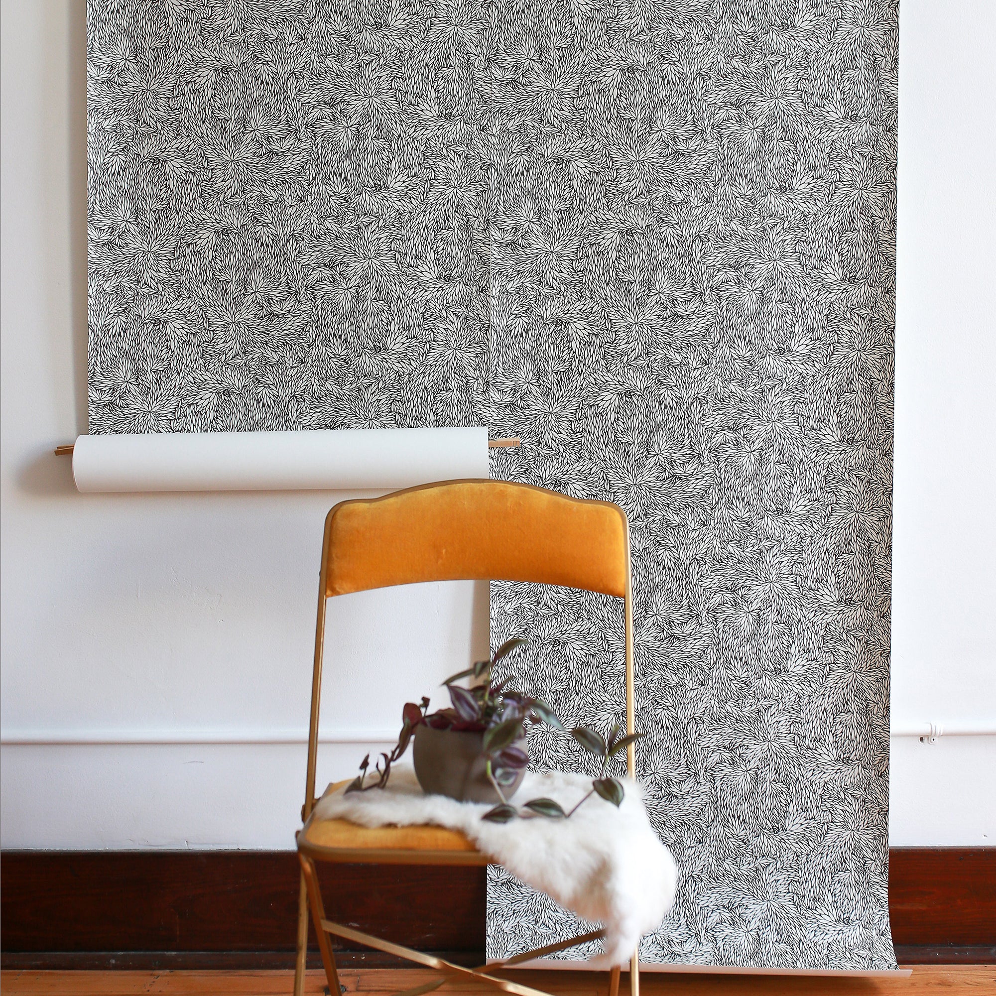  Interior space with a black and white botanical wallpaper roll partially hung on the wall, styled with a mustard velvet chair and a small potted plant.