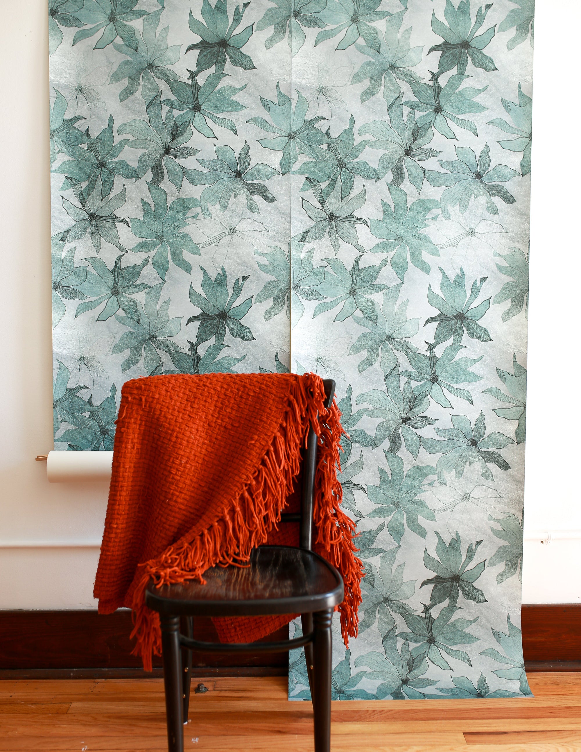 Light blue floral wallpaper displayed in hanging panels; black wooden chair with an orange woven throw draped over it in the foreground.