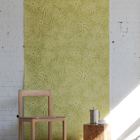 Minimalist room with light wooden chair, small square table, and stainless steel
coffee pot, set against green patterned wallpaper with swirling floral sun design.