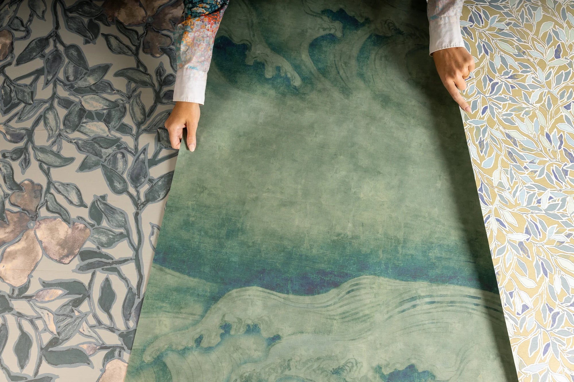 Elegant silk wallpaper samples being examined, featuring three distinct designs: a botanical pattern with dark green leaves on a muted taupe background, a painterly green wave-inspired design, and a delicate golden-yellow and blue leaf motif on a light background.