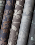 Close-up of six rolled botanical wallpaper designs in varying shades of gray, taupe, navy, and charcoal, each featuring a stylized leafy vine pattern with a matte finish.
