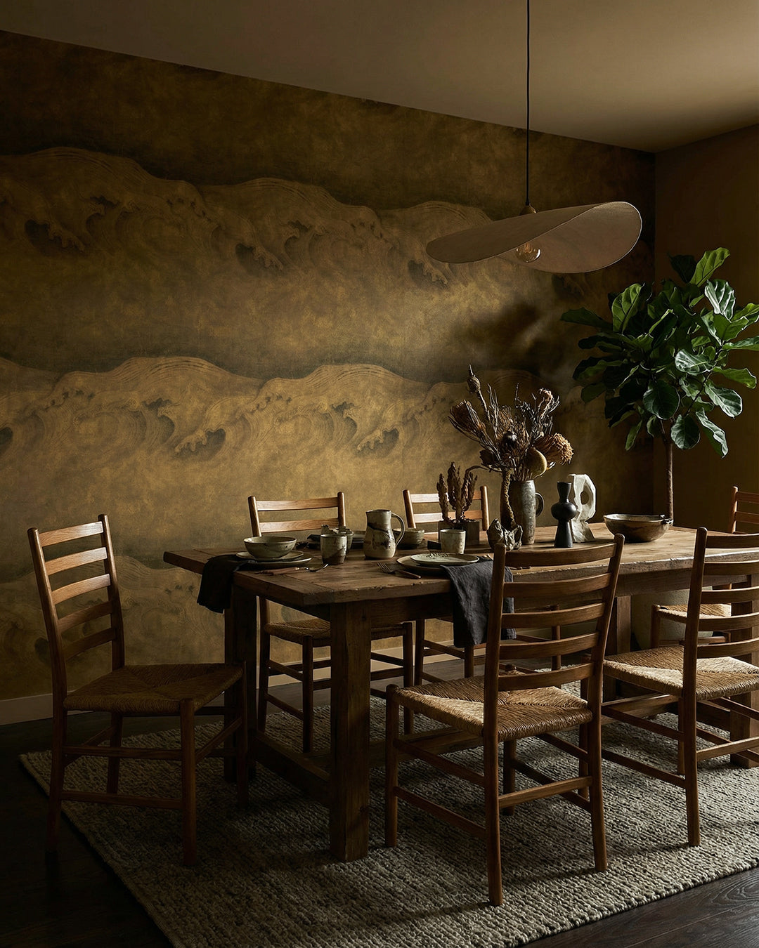 A dramatic, moody dining room featuring Wave Brass mural wallpaper with a textured golden wave motif, styled with a rustic wood farm table, rush-seat ladder-back chairs, dried botanical centerpiece, and a sculptural pendant light — Poppy wallpaper