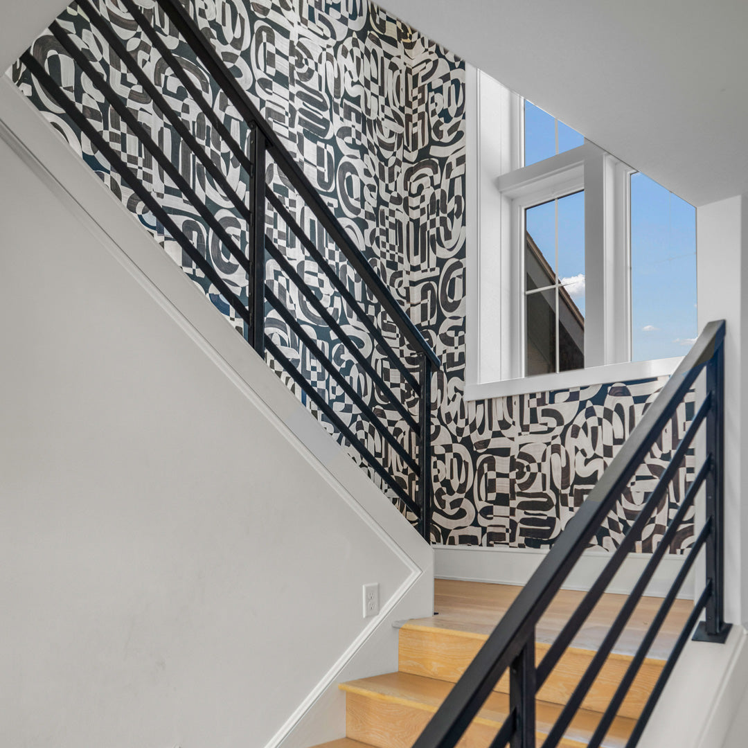 Modern stairwell featuring black-and-white grasscloth wallpaper with an abstract geometric pattern, paired with a black metal railing, light wood steps, and a large window allowing natural light.