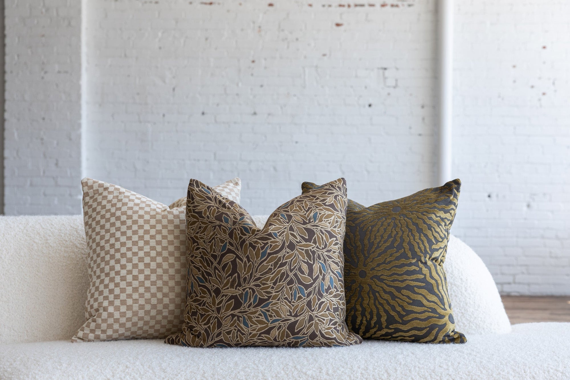 Dark brown with gold and blue botanical leaf pillow styled with cream checkerboard and gold sunburst pillows on white boucle sofa