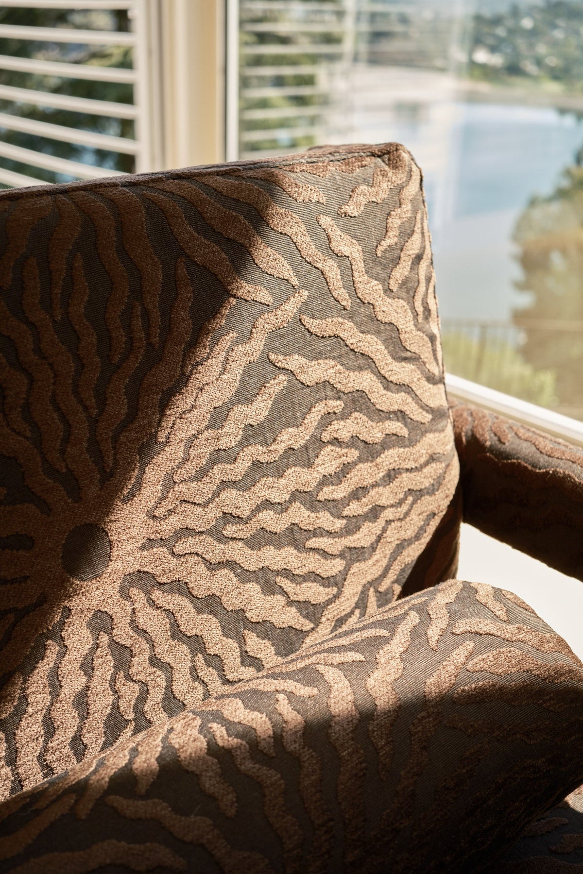 Brown sunburst jacquard fabric upholstered on a chair beside a sunlit shuttered window