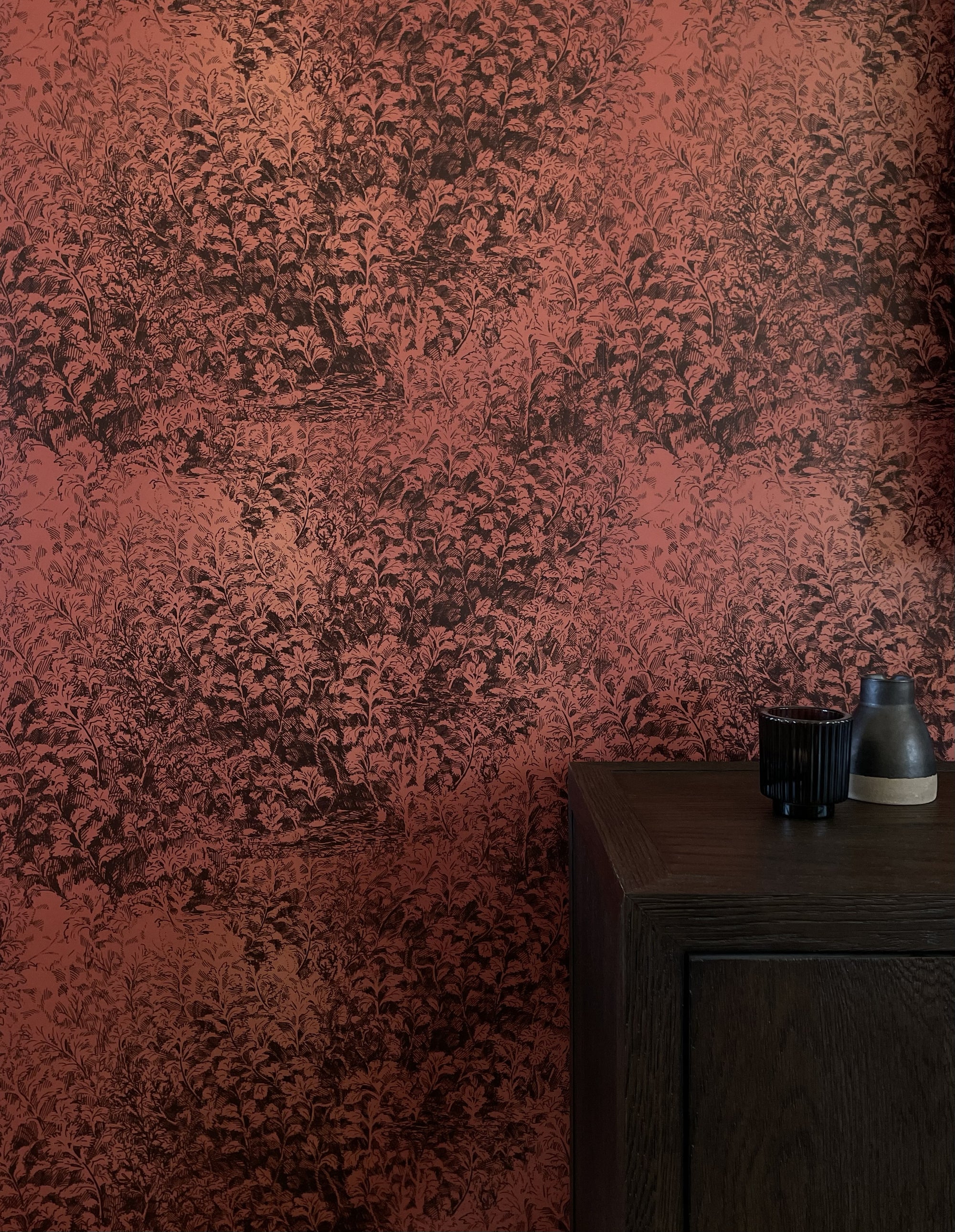 Dramatic botanical wallpaper in a deep rust-red hue with intricate foliage illustrations, styled with a dark wooden cabinet and minimalist decor.