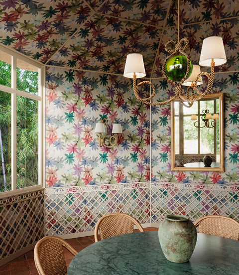 Dining room with multi-colored floral wallpaper covering walls and ceiling, featuring pink, green, and purple leaf patterns; green marble table, wicker chairs, and rustic chandelier with white shades in the foreground.